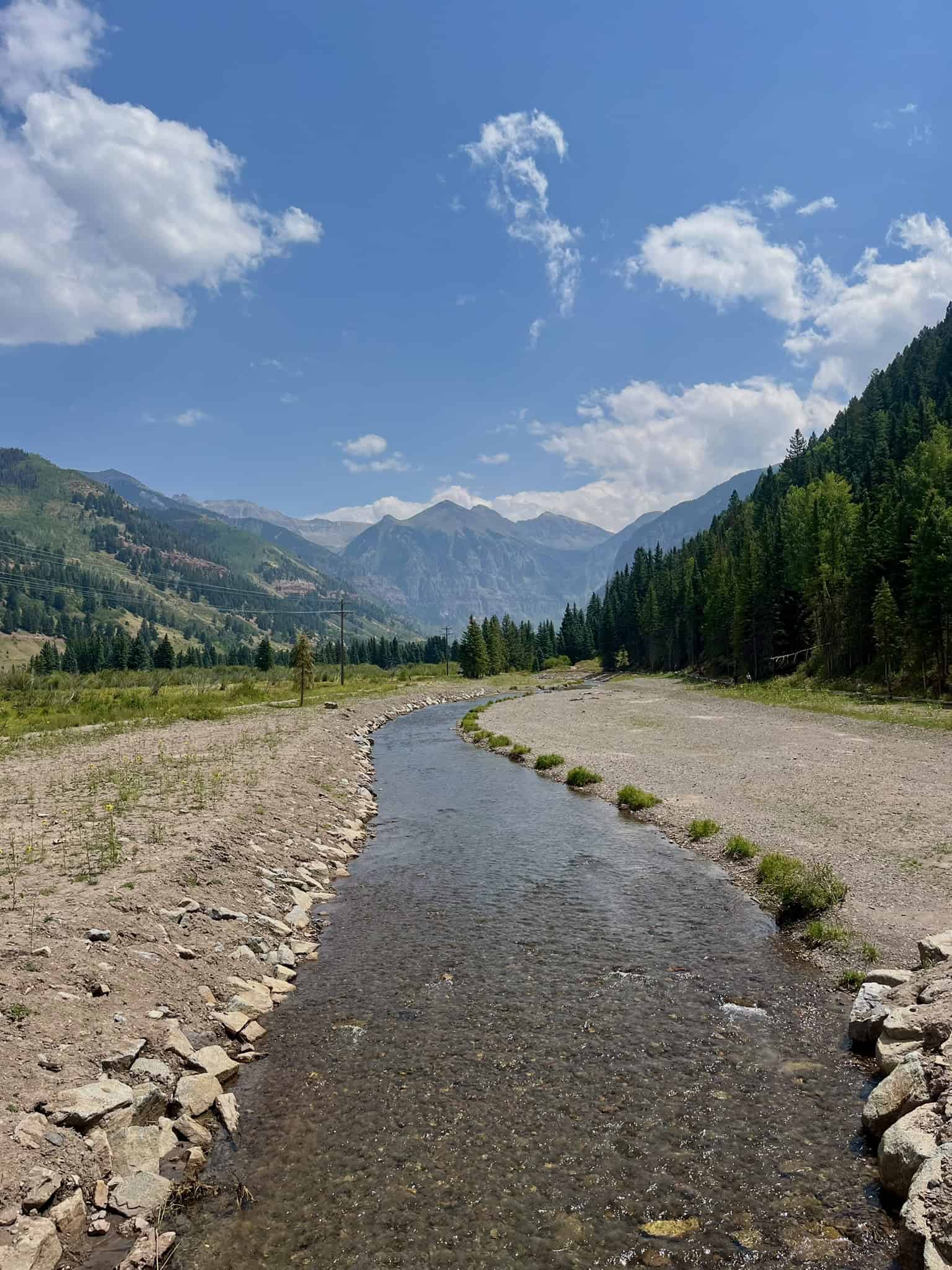 A wide river flows through a rocky valley floor, flanked by evergreen trees and leading toward the mountain peaks behind Telluride, Colorado.

