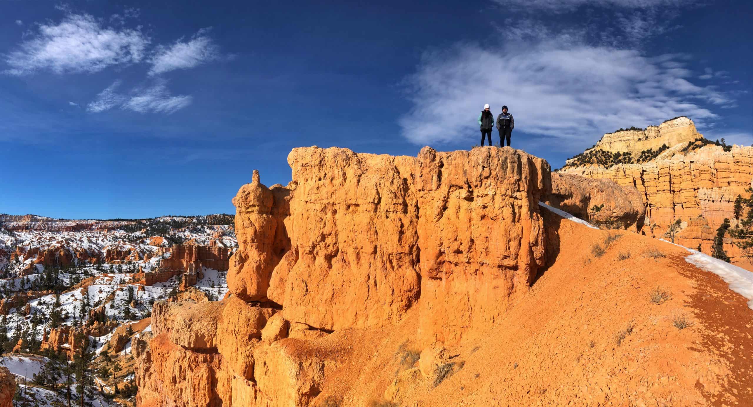 Two hikers on top of a Hoodoo on their one day Bryce Canyon Itinerary, enjoying the views of the snow dusted landscape under a blue sky