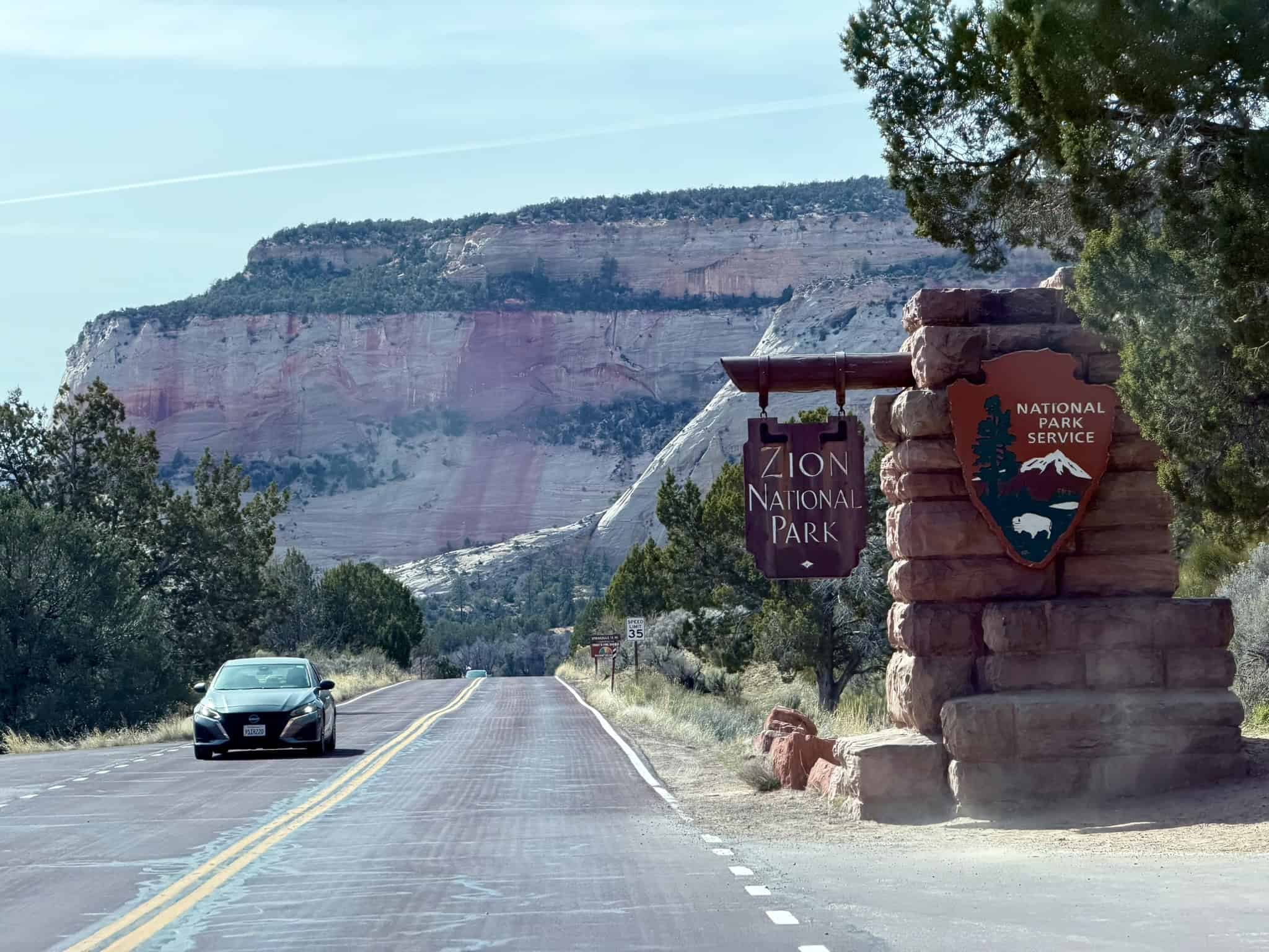 A vehicle travels down a paved road past a stone entrance monument toward towering red rock cliffs at Zion's National Park's east gate,