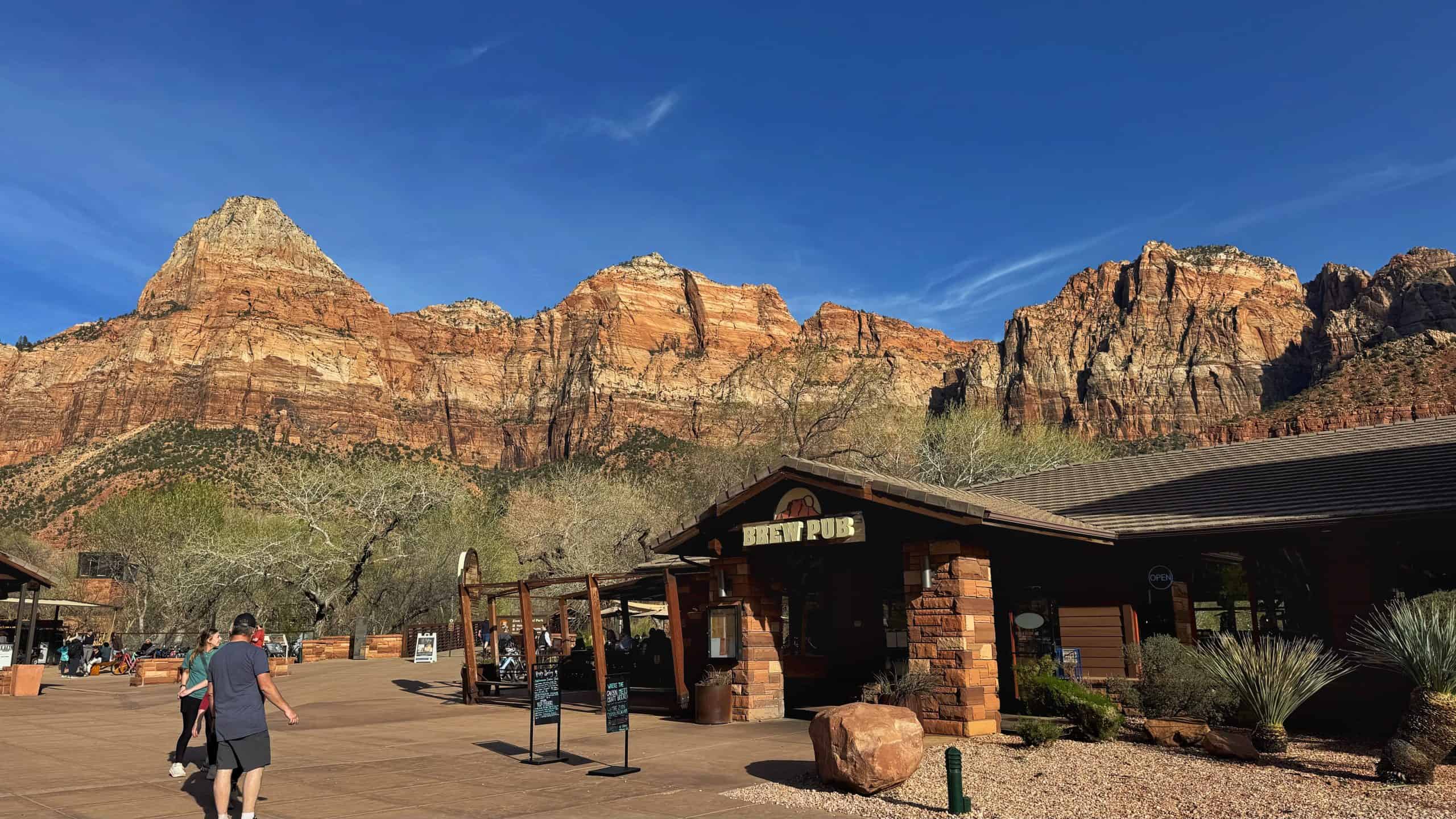 Visitors stroll past a brew pub at Zion Canyon Village, framed by towering red rock cliffs. making this area a key stop on any 2 day Zion national park itinerary.

