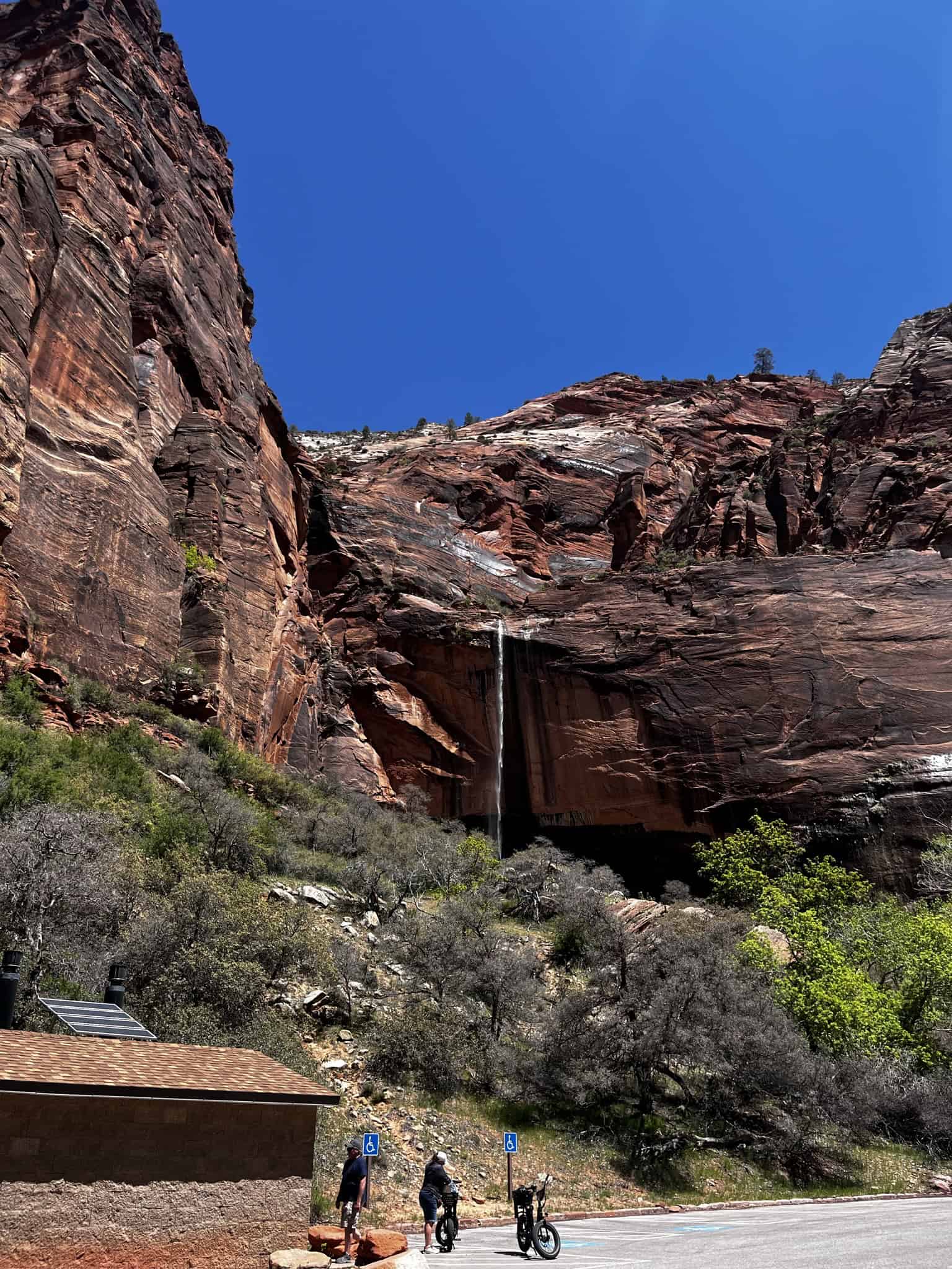 Visitors walk near a small building at the base of towering red cliffs where a seasonal waterfall cascades down at Weeping Rock in Zion National Park.