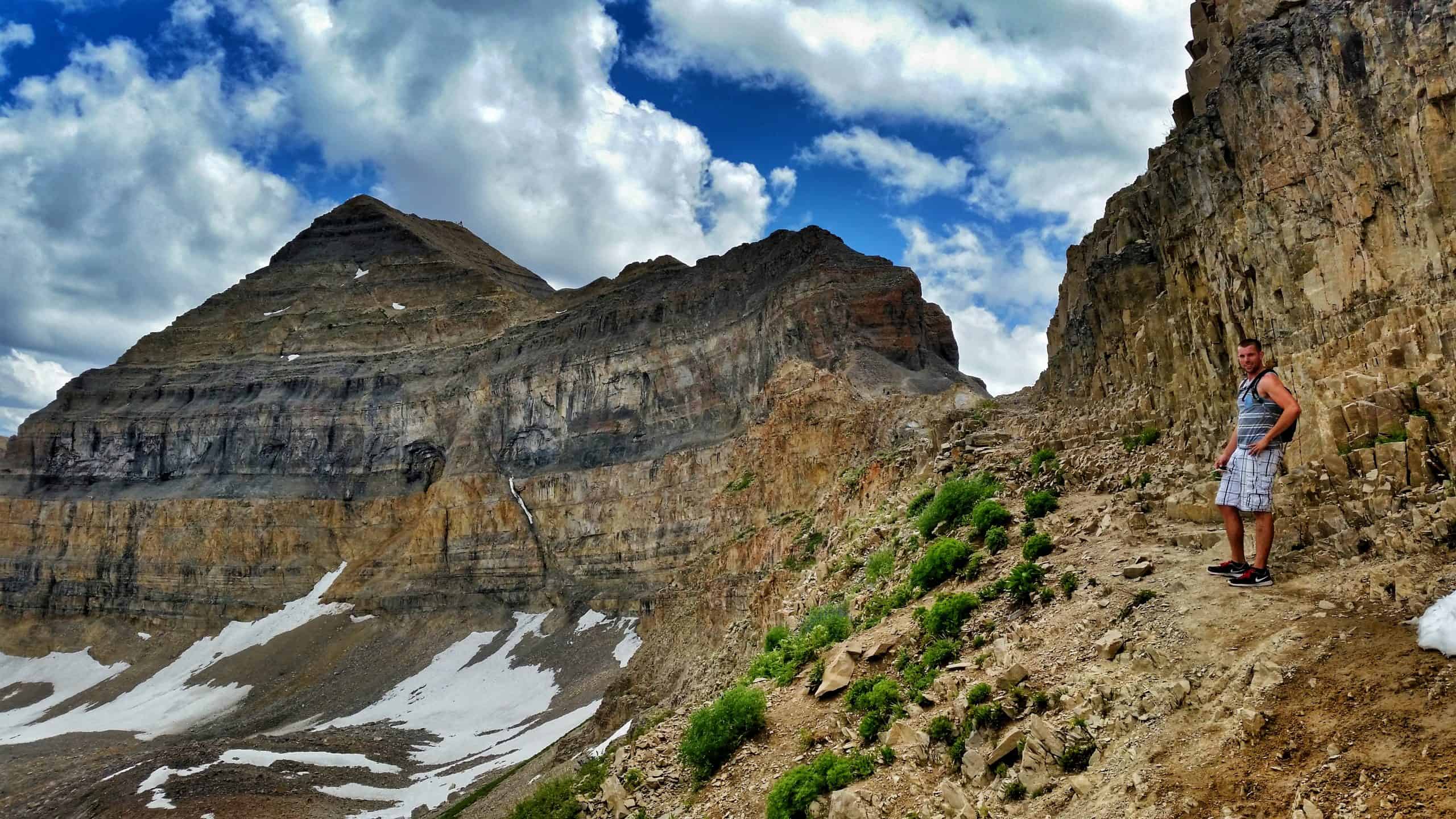 A hiker pauses on a narrow section of the Mount TImpanogos trail beside a steep cliff, with snowy slopes and a towering layered mountain rising above