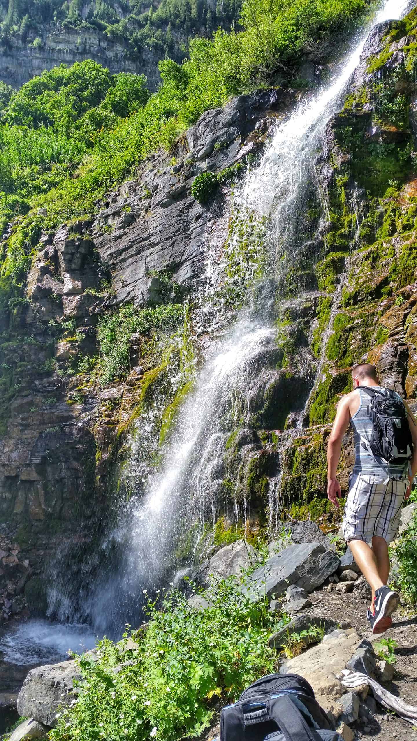 A hiker in a tank top and patterned shorts walks along the rocky terrain towards the rushing cascade of Second Falls on the Mount Timpanogos Hike.