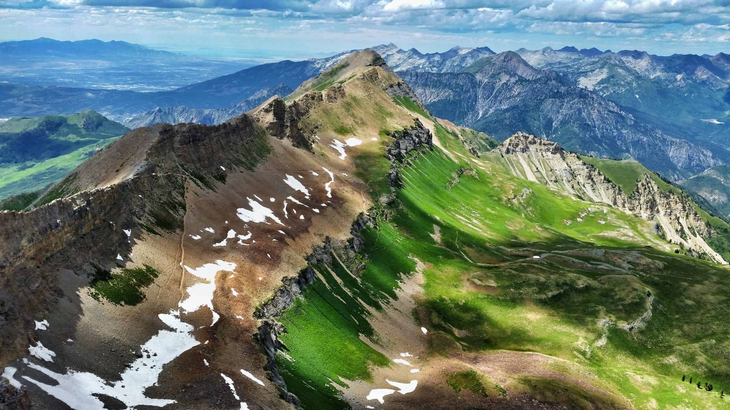 From the top of the Mount Timpanogos Hike, a rugged ridge of vibrant green slopes and lingering snow stretches towards layered mountain ranges in the background under a bright, partly cloudy sky.