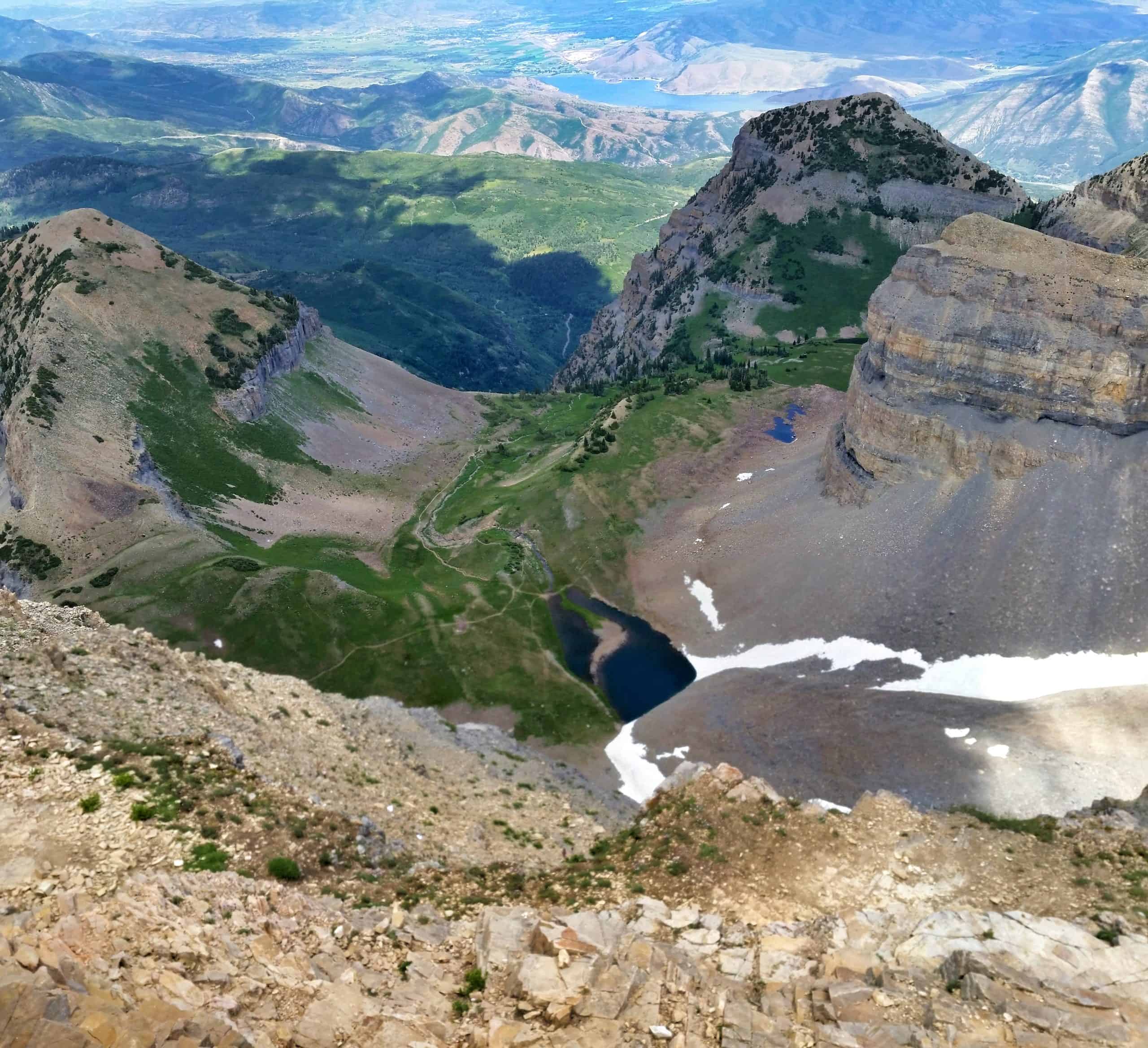 A dramatic vista looking down from the summit of Mount Timpanogos with a seasonal snowfield on a rocky slope leading towards a blue lake, meadows and distant mountain peaks.