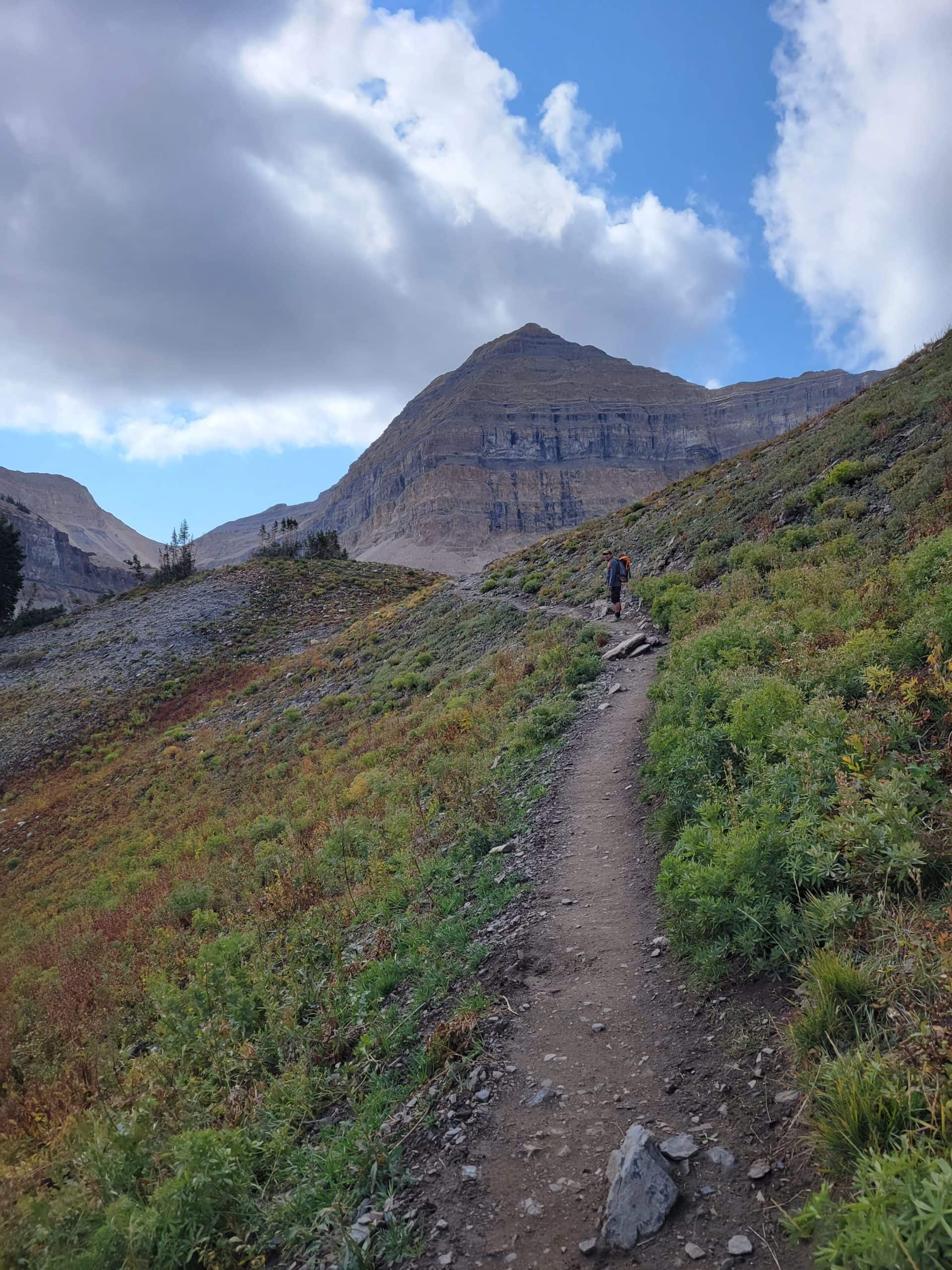 A narrow dirt trail climbs towards an ascending hiker beneath a dramatic, layered peak on the Mount Timpanogos Trail.