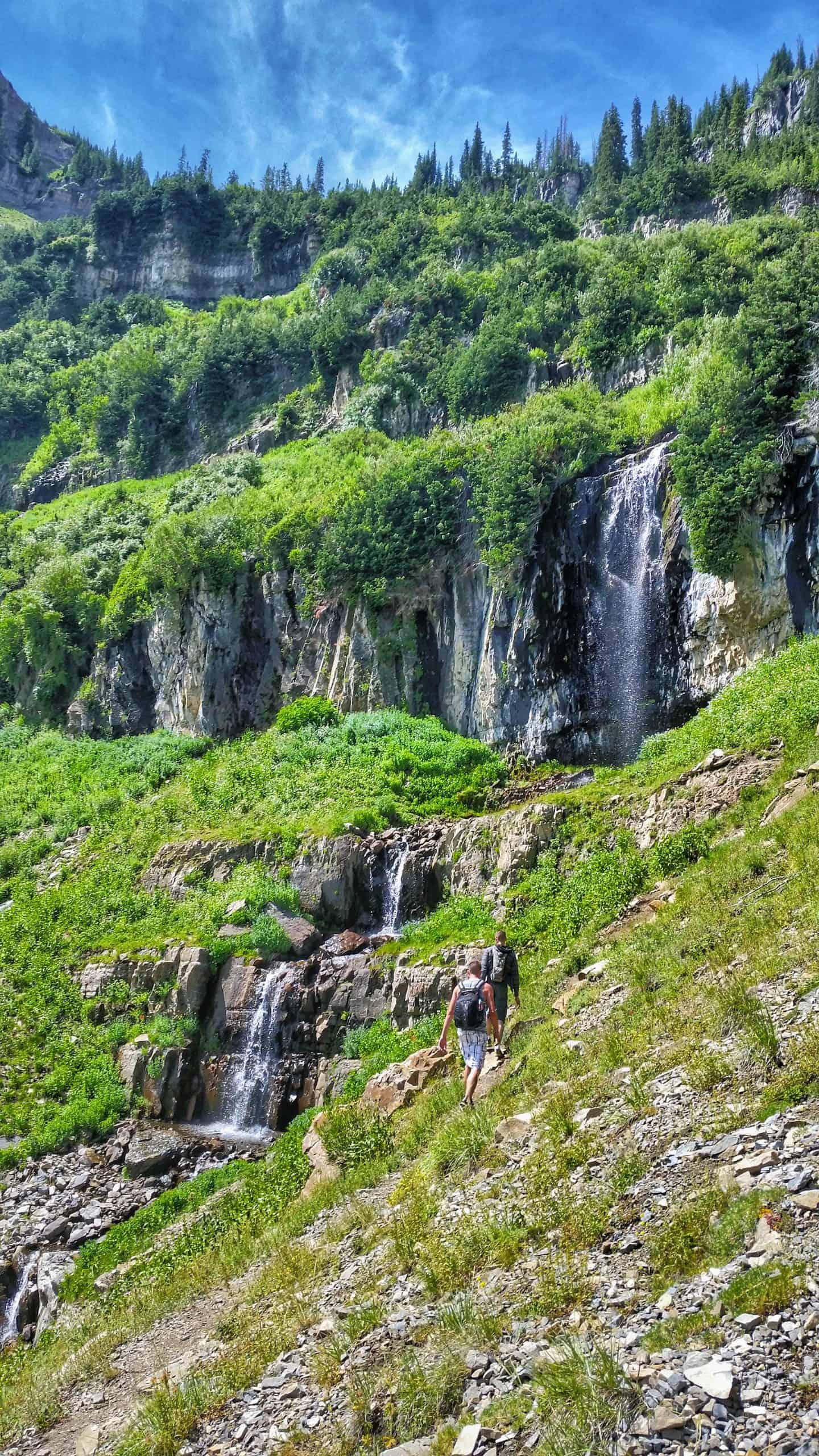 Hikers walking along a grassy, rocky incline, towards several cascades tumbling down the rugged cliff face on the Mount Timpanogos Hike.