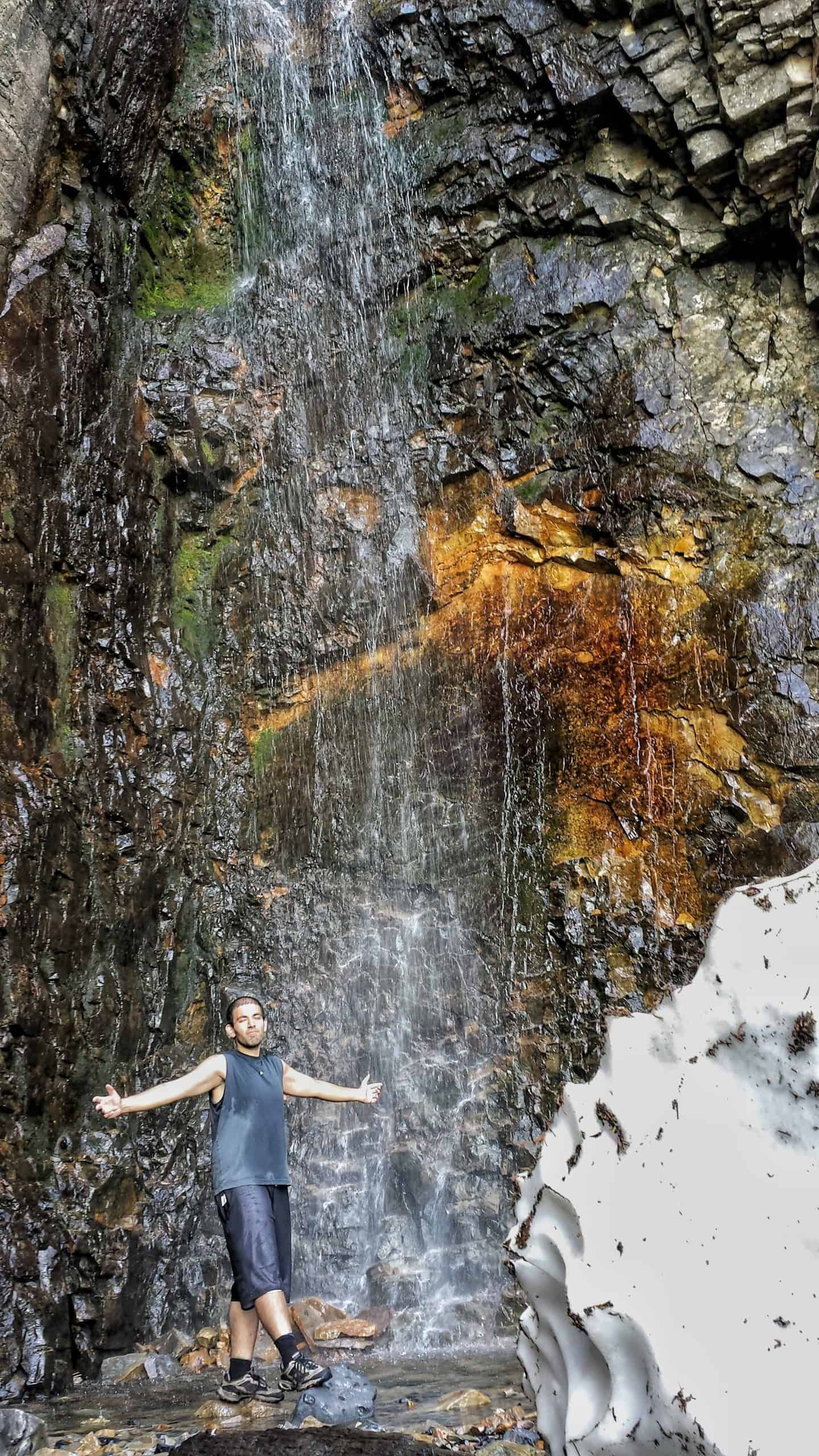 A hiker stands beside a waterfall on the Mount Timpanogos Hike, as it flows over a colorful rock face, with a patch of lingering snow visible to the side.