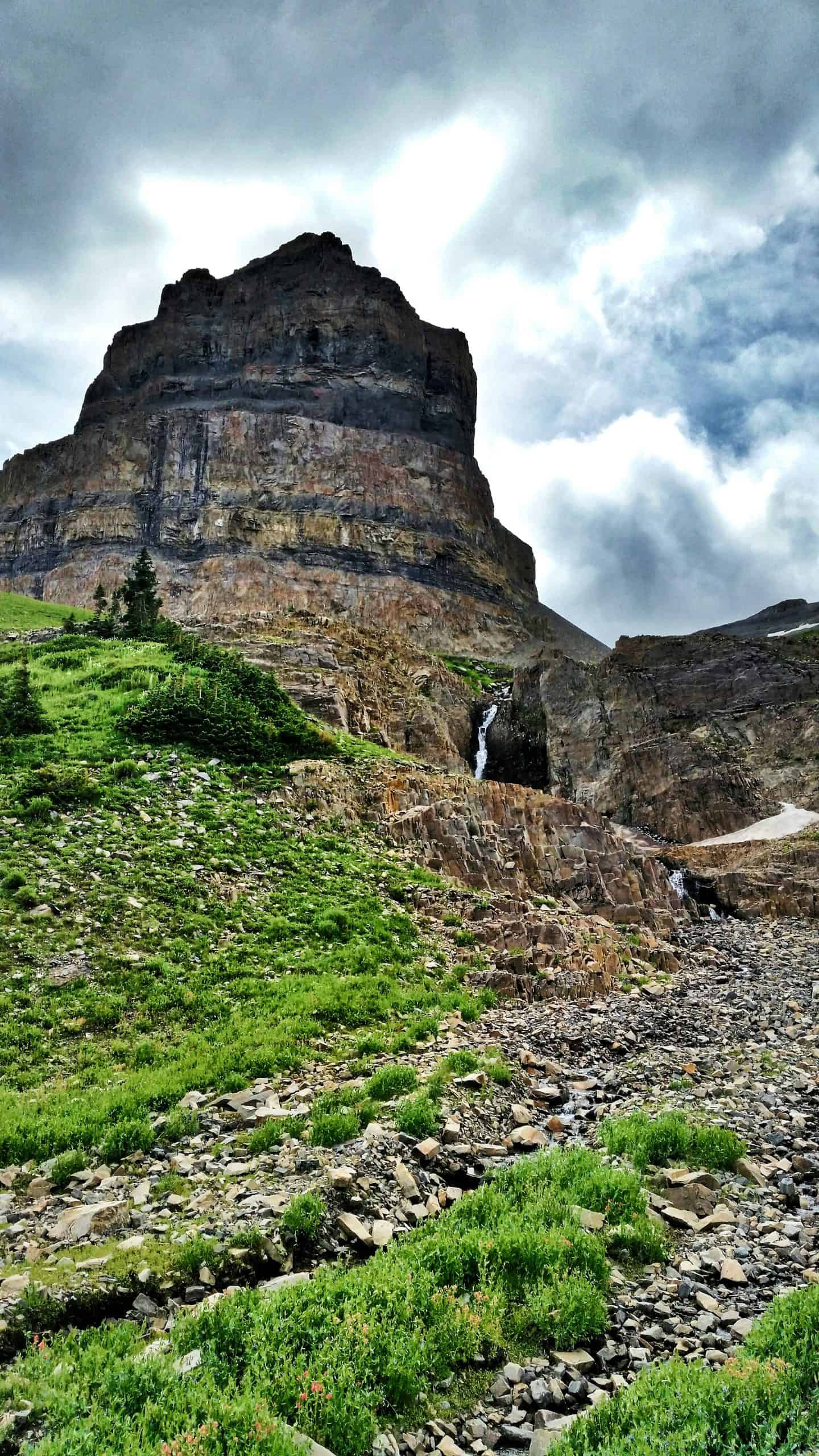 A waterfall tumbles down a narrow vertical crack on the Mount Timpanogos hike, with a massive, layered mountain the background, beneath a cloudy sky