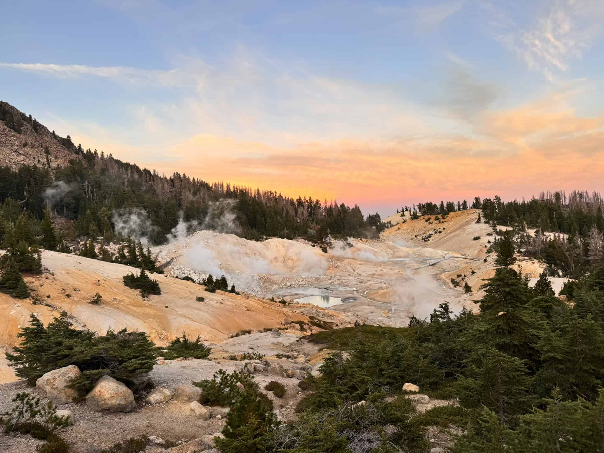 A geothermal landscape at Bumpass Hell in Lassen Volcanic National Park, featuring steaming vents and vibrant mineral terraces under a warm, golden sky.