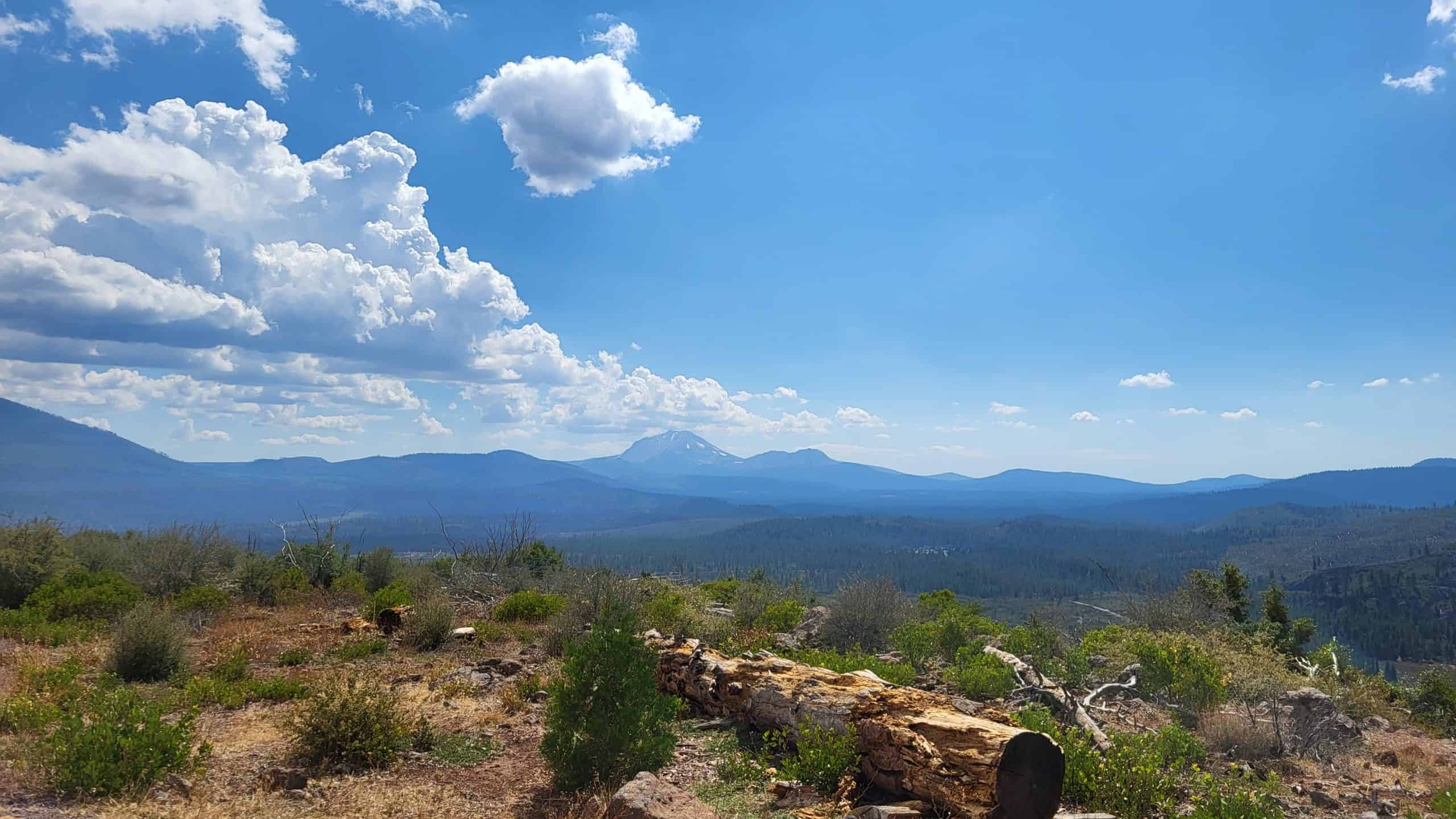 Lassen Peak rises above layered blue mountain ridges and forested slopes, viewed from the Hat Creek Rim Scenic Viewpoint under a partly cloudy sky.