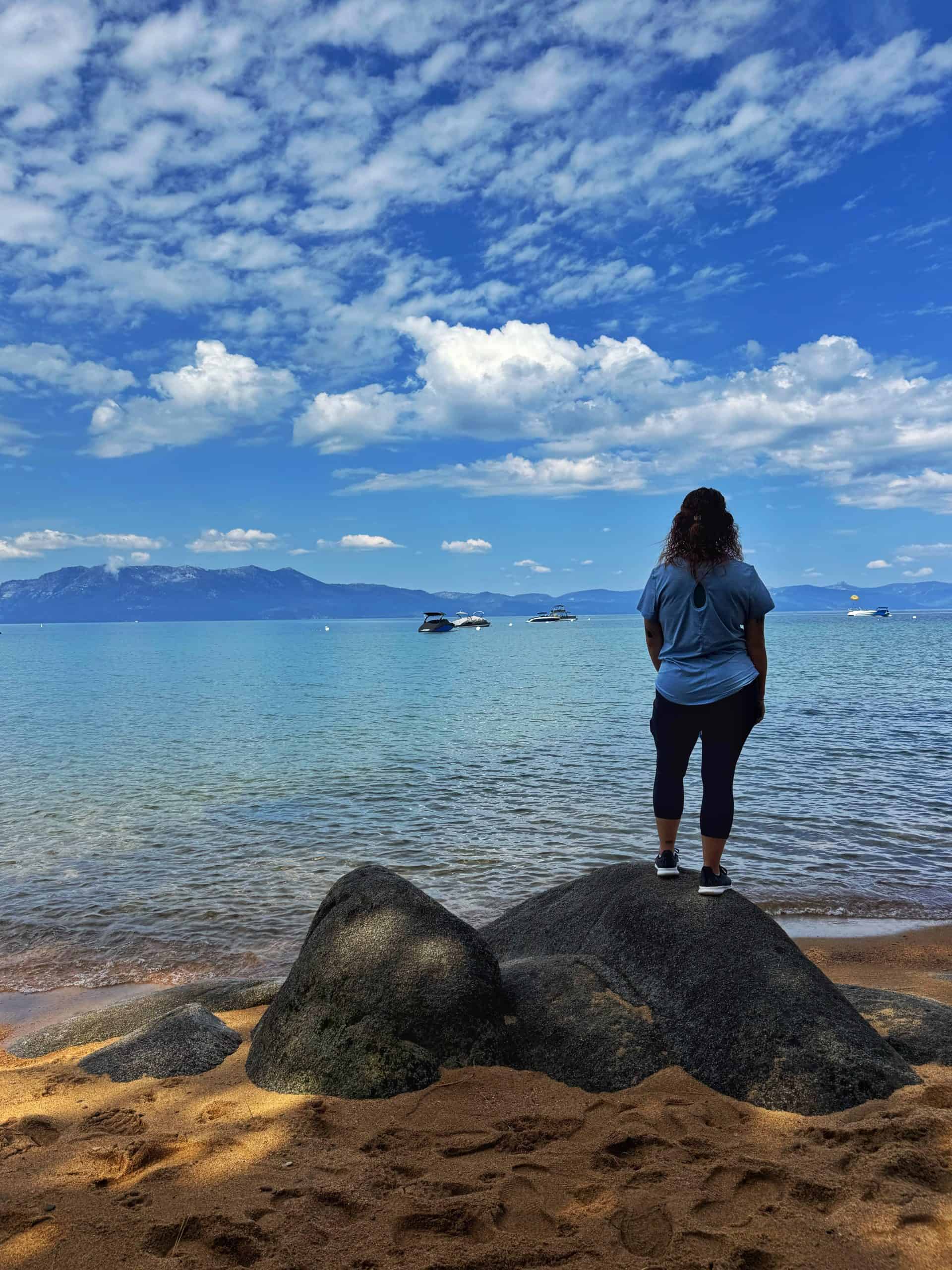 A Woman stands on a boulder along the sandy shore of Zephyr Cove, Lake Tahoe, gazing out at the clear blue water and distant mountains under a beautiful sky.