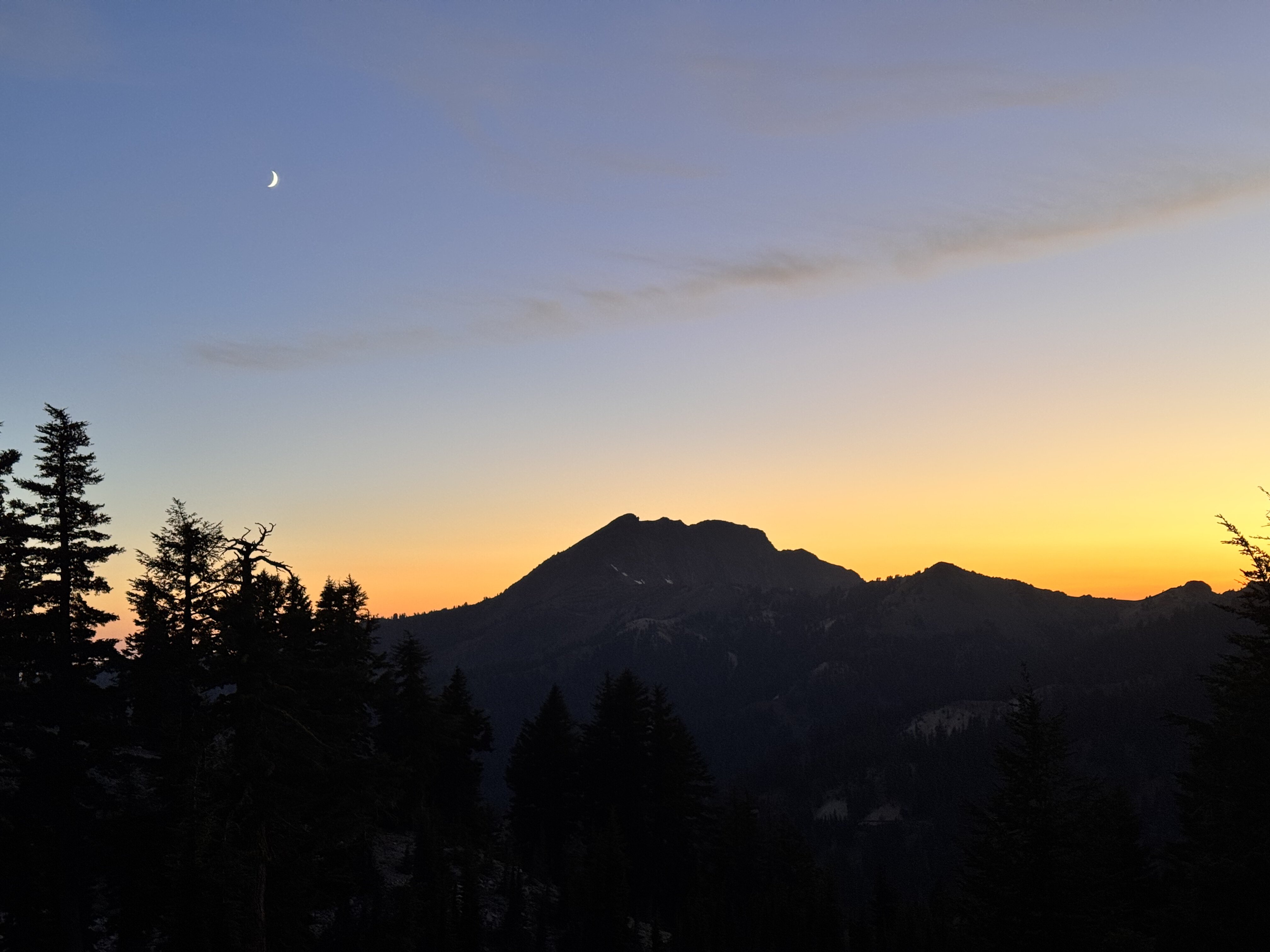 A silhouette of Brokeoff Mountain and pine trees under a golden hour sky and a crescent moon, as seen from the Bumpass Hell trail