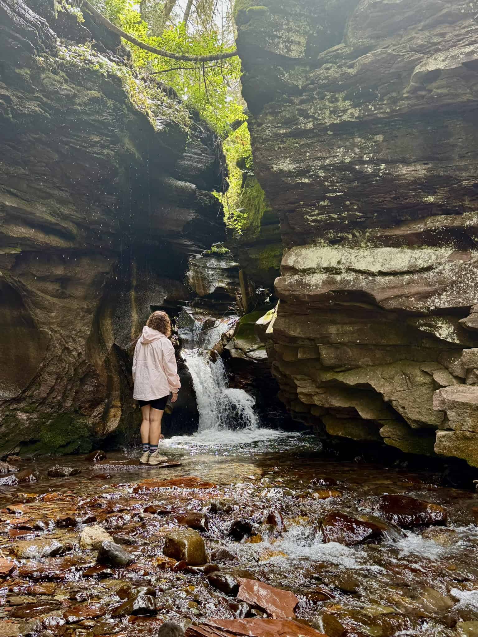 A hiker stands on wet rocks beneath a waterfall emerging from a rocky cave, capturing the scenery of the Little Hawaii hike in Telluride.