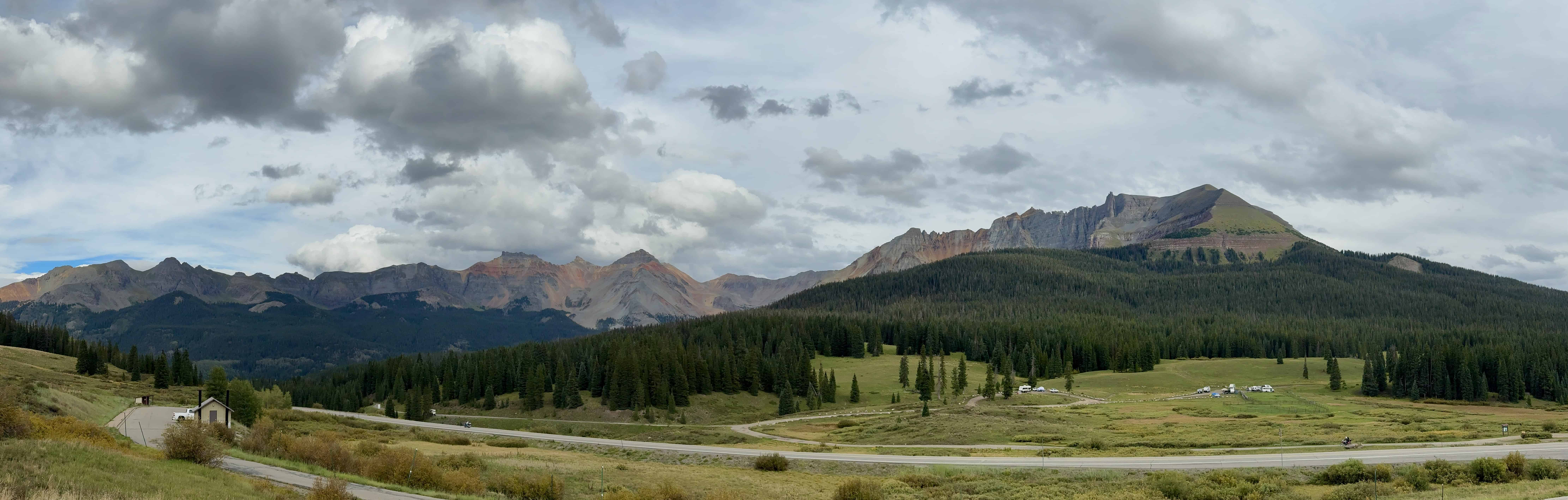 A winding road cuts through a lush green valley surrounded by forests and rugged mountain as seen from Lizard Head Pass near Telluride,
