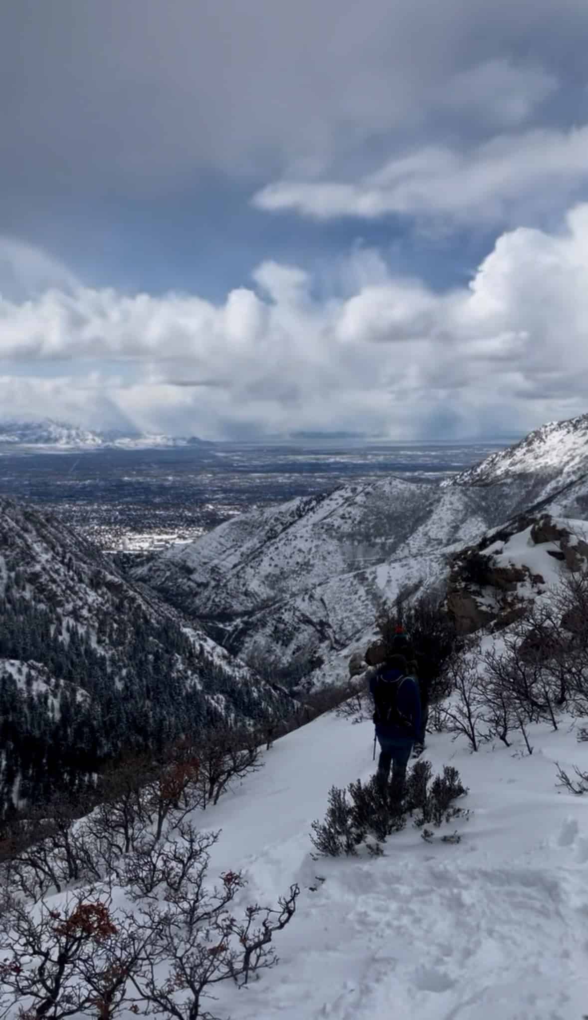 A hiker with a backpack walks along a snowy mountain ridge, overlooking a wide valley and mountains in the distance under a cloudy sky.