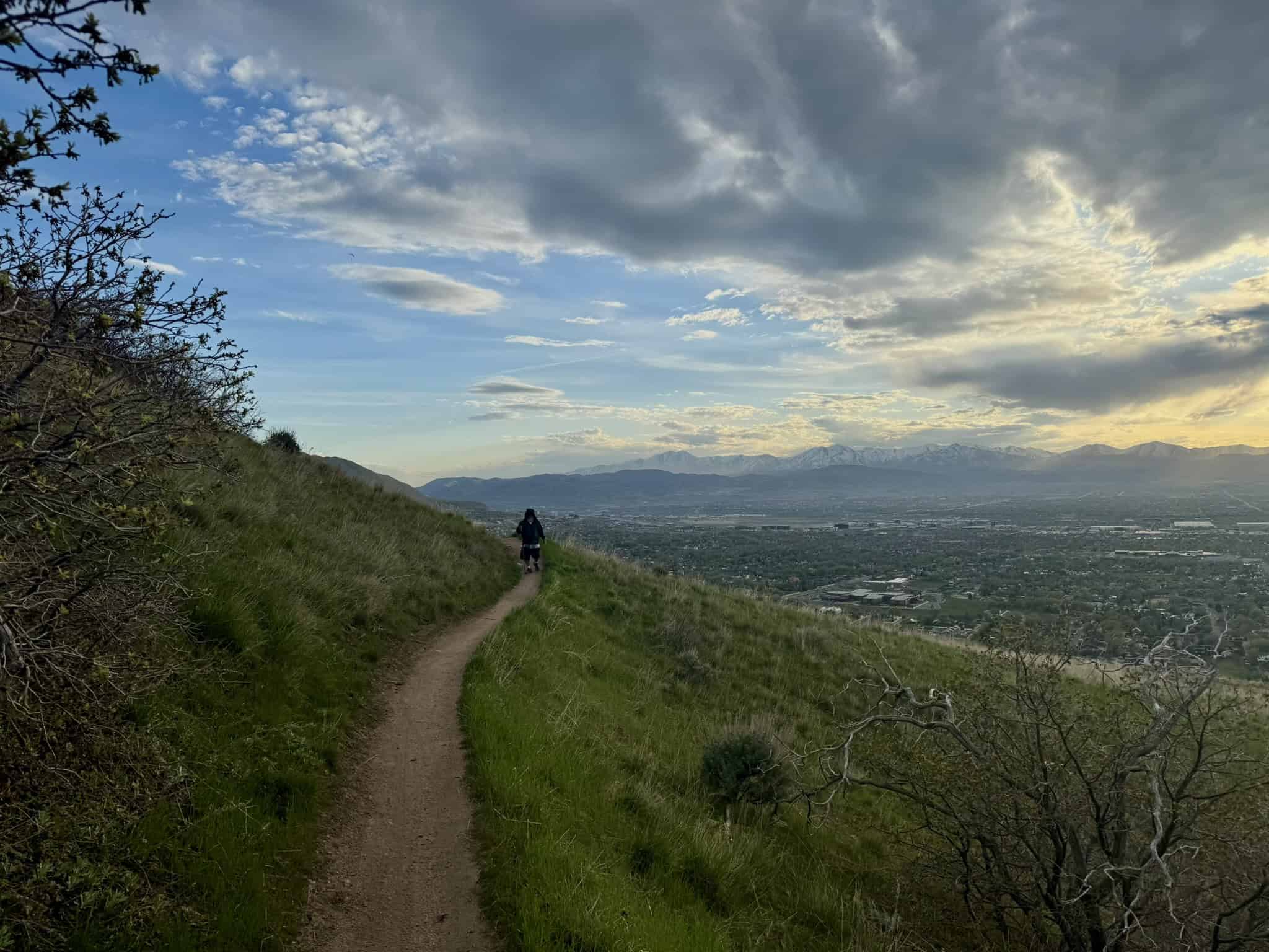Hiker walking along a hillside trail above the Salt Lake Valley beneath dramatic clouds and mountain views